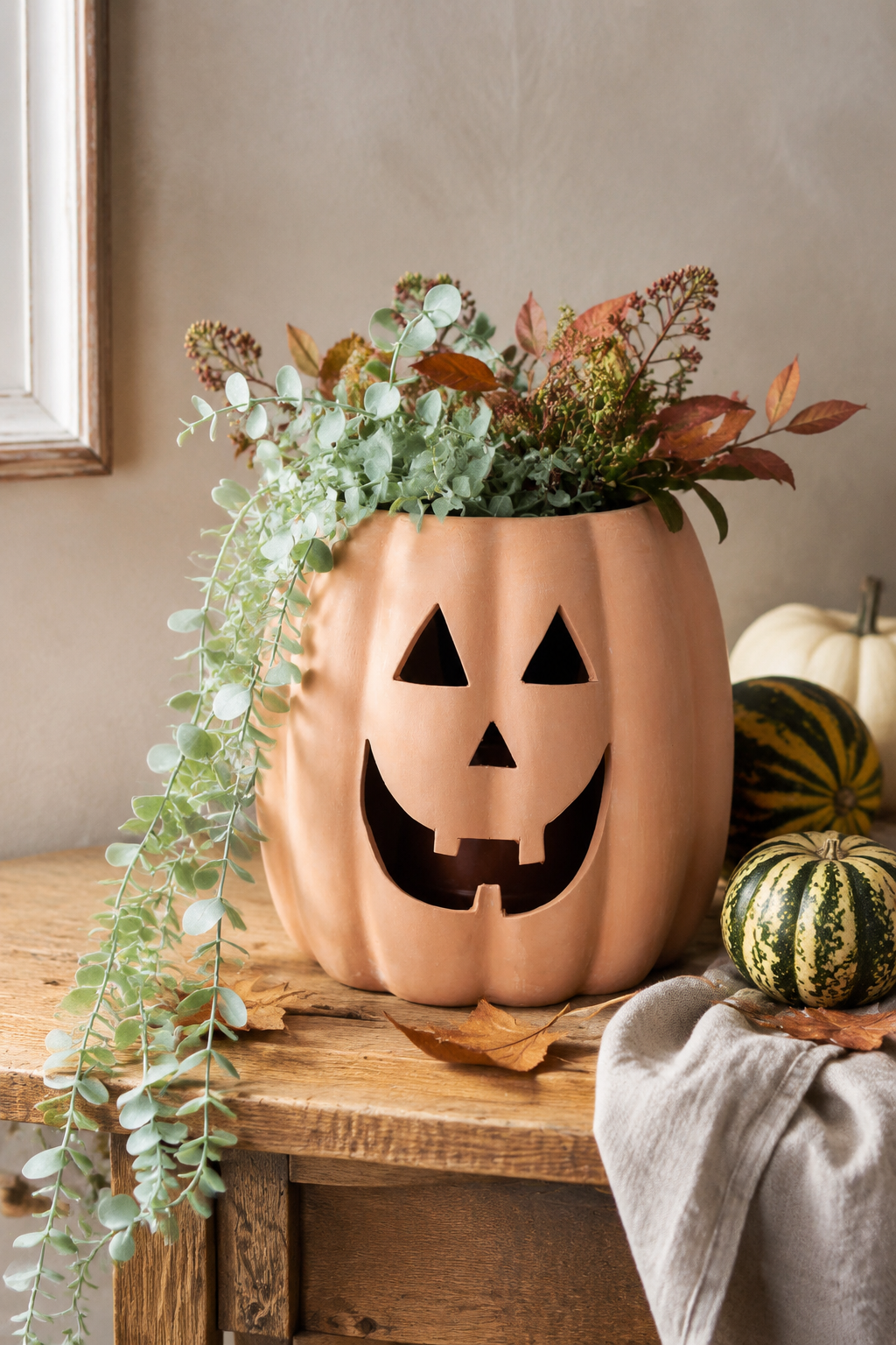 A terracotta-colored pumpkin-shaped planter with a carved jack-o'-lantern face, filled with green eucalyptus and autumn foliage, displayed on a rustic wooden table alongside small gourds and a draped fabric.