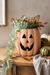 A terracotta-colored pumpkin-shaped planter with a carved jack-o'-lantern face, filled with green eucalyptus and autumn foliage, displayed on a rustic wooden table alongside small gourds and a draped fabric.