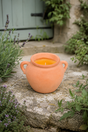 An orange terracotta clay candle holder with two side handles, filled with a yellow-orange citronella candle, set on a stone ledge outdoors surrounded by green plants and lavender.