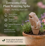 Terracotta frog plant watering spike with plant in a pot, set against a garden background.