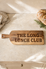 A rustic wooden serving board with the phrase 'THE LONG LUNCH CLUB' engraved in dark lettering, styled on a light stone countertop with bread and a cloth nearby.