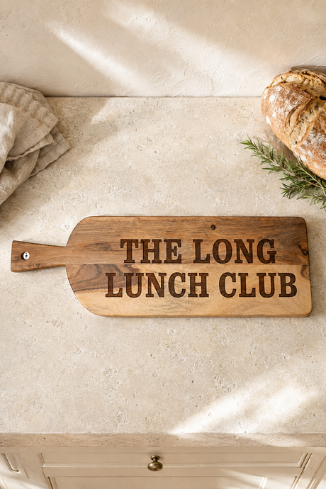 A rustic wooden serving board with the phrase 'THE LONG LUNCH CLUB' engraved in dark lettering, styled on a light stone countertop with bread and a cloth nearby.
