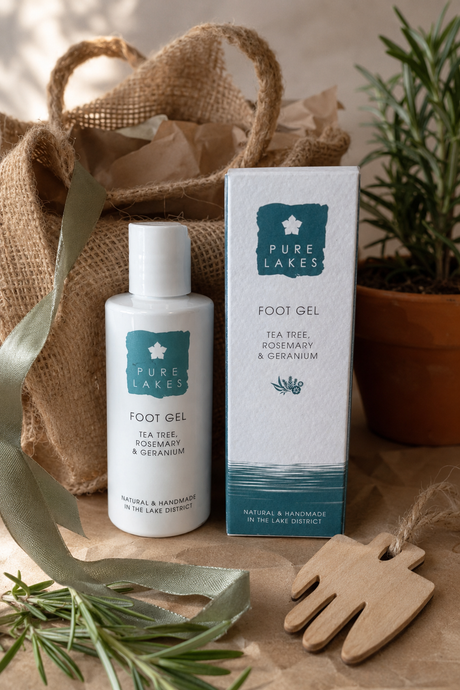 Pure Lakes Foot Gel bottle and packaging box displayed on a rustic brown surface with a burlap bag, ribbon, rosemary sprigs, and a wooden hand-shaped object in the background.