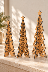 Three laser-cut wooden Christmas trees in different heights, arranged on a light stone surface against a warm beige wall with soft sunlight and shadows.