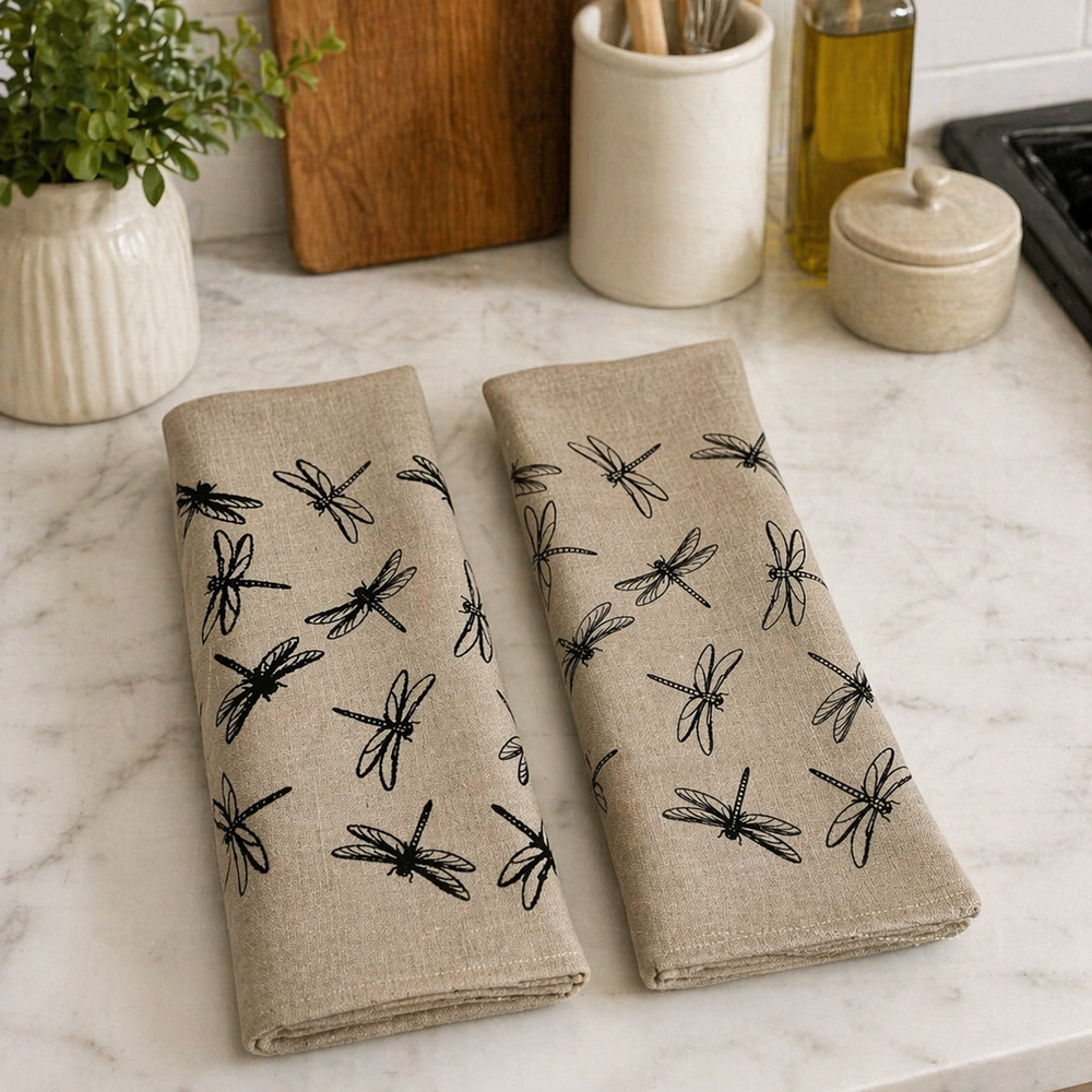Two beige linen tea towels with black dragonfly patterns on a kitchen counter.