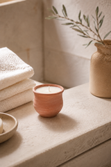 terracotta pot candle on a stone surface, next to a stack of towels and a vase with greenery.