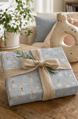 A gift box wrapped in light Vintage Fashion Sketch Wrapping Paper Pastel Blue, tied with a rustic burlap ribbon and a sprig of green pine, set on a wooden table in a cozy living room.