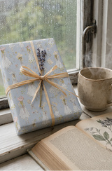 A wrapped gift box with aVintage Fashion Sketch Wrapping Paper Pastel Blue, tied with a raffia ribbon. Placed on a rustic windowsill next to an open book and a ceramic mug. The gift box is tied with a lavender sprig.