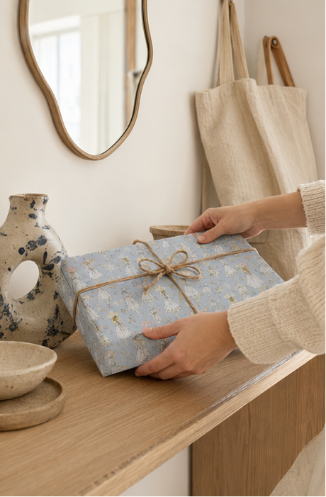 A person's hands are shown holding a gift box wrapped in Vintage Fashion Sketch Wrapping Paper in Pastel Blue and tied with a rustic brown twine bow. The box is placed on a light wood console table in a cozy, neutral-toned room with a speckled vase, a canvas tote bag, and a mirror in the background.
