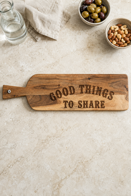 A rustic wooden serving board with the engraved phrase 'GOOD THINGS TO SHARE' in dark brown lettering, styled on a light beige stone surface with bowls of olives and nuts nearby.