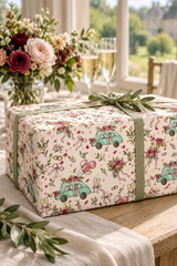 A square gift box wrapped in Wacky Wedding Car Wrapping Paper in Mint Green & Pink, tied with a sage green ribbon, set on a wooden table in a bright, sunlit room with flowers and champagne glasses in the background.