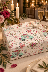 A gift box wrapped in white paper printed with a romantic wedding-themed pattern of mint green cars, pink roses, hearts, and champagne glasses, tied with a white satin ribbon. The box is set on a decorated table with lit candles, flowers, and champagne glasses in the background.