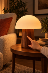 Hand touching the copper control button on a large walnut Alice Mushroom Lamp beside a cream armchair