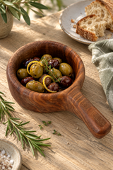 A wooden bowl filled with stuffed green and black olives, garnished with lemon slices and herbs, set on a rustic wooden table with bread, rosemary, and a napkin nearby.