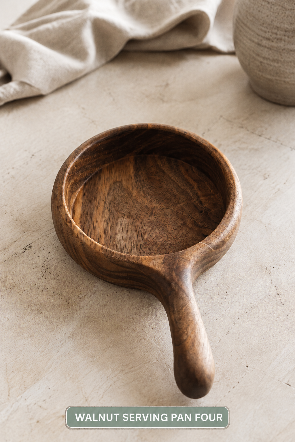 A walnut serving pan with a rich dark brown wood grain, shown on a neutral textured beige surface with a linen cloth and ceramic vase in the background. A text label at the bottom reads 'WALNUT SERVING PAN FOUR'.