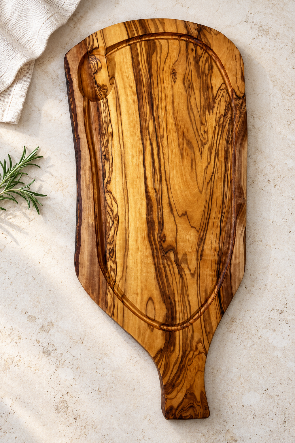 An olive wood serving board with a decorative handle and a built-in loop, photographed from above on a light stone surface with a linen cloth and rosemary sprig as props.