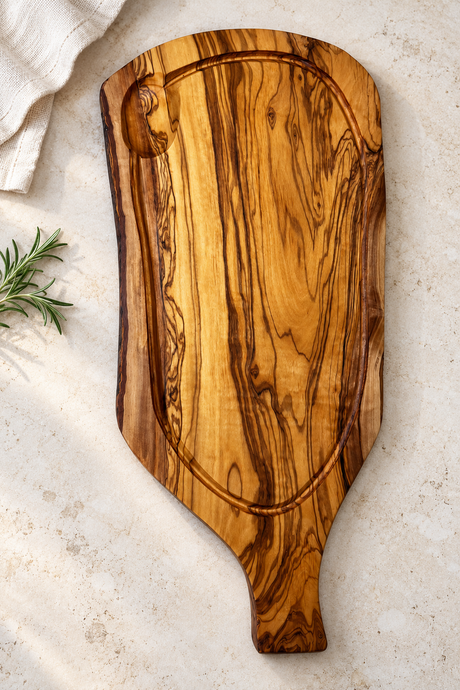 An olive wood serving board with a decorative handle and a built-in loop, photographed from above on a light stone surface with a linen cloth and rosemary sprig as props.