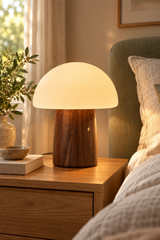 Large walnut Alice Mushroom Lamp glowing warm white on a bedside table beside linen bedding.