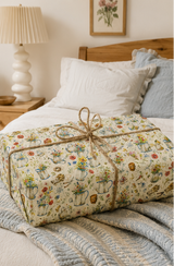A wrapped gift box tied with a jute twine bow, covered in a vintage floral and botanical pattern with watering cans, flowers, and garden tools on a cream background, resting on a cozy bed with white and blue pillows in a warm bedroom setting.