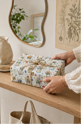 A person's hands are shown holding a wrapped gift box with floral-patterned wrapping paper with watering cans on it in a vintage style. The gift is tied with a twine bow, set on a wooden shelf in a cozy, neutral-toned room.