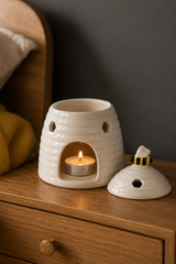 A ceramic beehive-shaped tea light candle warmer with a lit tea light inside, shown on a wooden bedside table next to its matching bee-shaped lid against a dark gray wall.