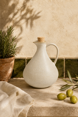 A matte ceramic olive oil dispenser with a cork stopper, styled on a stone surface with olives, a potted plant, and a textured wall in warm natural light.