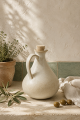 A matte ceramic olive oil bottle with a cork stopper, styled on a stone surface with olive branches, olives, a potted plant, and draped fabric against a textured wall with leaf shadows.