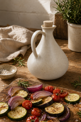 A matte off-white ceramic oil bottle with a cork stopper sits on a rustic wooden table next to a cutting board with roasted zucchini, red onion, and cherry tomatoes. A small bowl of salt and a potted rosemary plant are visible in the background.