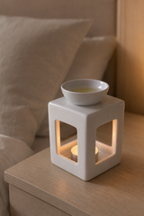 A white ceramic cube-shaped candle warmer with a small top bowl and glowing tea light candle inside, placed on a wooden bedside table next to a bed.