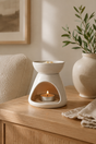 White ceramic oil diffuser with a lit candle on a wooden surface, next to a vase with greenery.