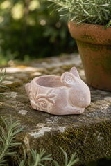 A small pale pink bunny-shaped Whitewashed Terracotta Rabbit Tealight Holder – 9.5cm with carved floral details, placed on a mossy stone surface outdoors with a potted plant in a blurred garden background.