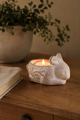 A small sculpted candle holder shaped like a rabbit, filled with a lit tealight candle, placed on a wooden table with a blurred potted plant and book in the background.