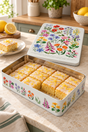 A decorative metal tin filled with small yellow sponge cake squares topped with white icing and yellow zest, set on a kitchen counter. The tin and its lid are decorated with colorful wildflower illustrations, and lemons, a plate, and a fork are visible nearby.