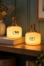 Mochi Wonder and Peek lamps glowing on a wooden shelf against a blue-green panelled wall.