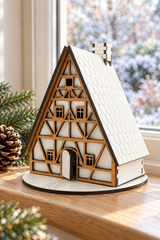 A miniature wooden half-timber house model with a white textured roof sits on a wooden windowsill, decorated with pine branches and a pinecone. A blurred snowy outdoor scene is visible in the background.