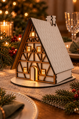 A miniature wooden A-frame house ornament with a white shingled roof, dark brown timber framing, and glowing warm yellow windows, displayed on a holiday table runner with pine branches and candles in the background.