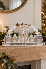 A decorative white Christmas villagescape tabletop ornament with a church spire, small houses, trees, reindeer, and a base reading 'Merry Christmas and Happy New Year,' styled on a wooden mantel with candles, pine branches, and warm string lights in the background.