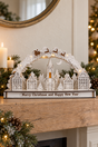 A decorative white Christmas villagescape tabletop ornament with a church spire, small houses, trees, reindeer, and a base reading 'Merry Christmas and Happy New Year,' styled on a wooden mantel with candles, pine branches, and warm string lights in the background.
