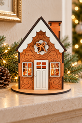 A miniature wooden gingerbread house ornament with carved details, white trim, warm glowing windows, and a small chimney, displayed on a tabletop with holiday greenery and blurred lights in the background.