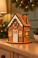 A decorative miniature house ornament with carved wood details, a warm glowing light inside, and a round wooden base, displayed on a wooden cabinet in a holiday setting with pine branches and warm lights.