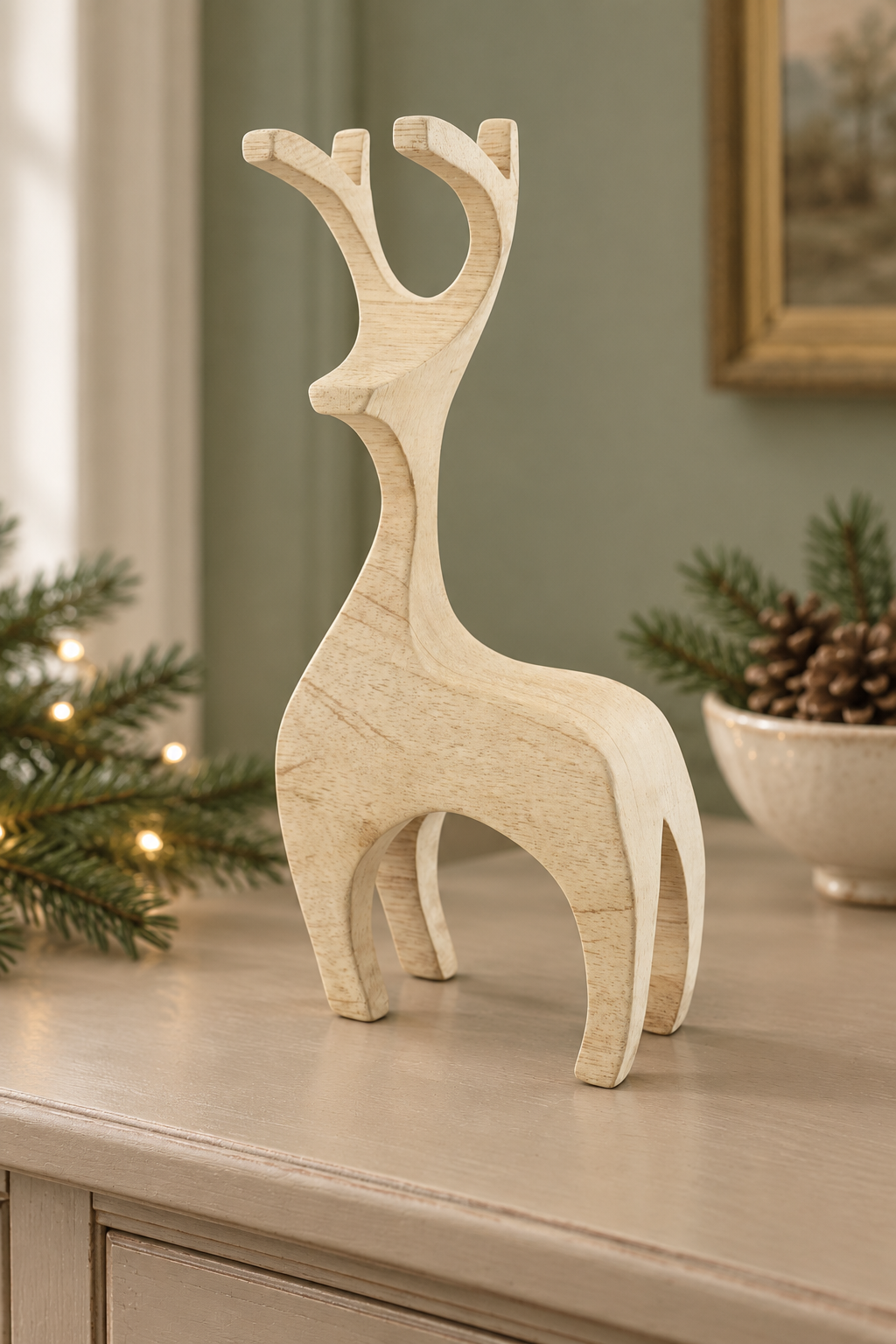 A light natural wood reindeer figurine standing on a tabletop in a styled indoor setting with holiday greenery, a bowl of pinecones, and a muted green wall in the background.