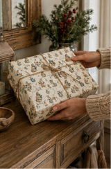 Illustrated woodland animal Christmas wrapping paper featuring scarf-wearing foxes, rabbits and mice sharing hot drinks and mince pies at a winter garden party. The pencil-sketch design includes lanterns, wrought-iron tables, watering cans, holly and snowy branches in teal, russet, pink, mustard, red and green on warm ivory.