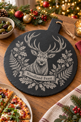 A round black slate serving board with a carved stag and the words 'Woodland Feast,' styled on a wooden table with holiday greenery, ornaments, a bowl of olives, pizza, and a candle.