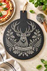 A round black slate cutting board with a hanging loop, featuring a detailed engraving of a stag with antlers and the words 'Woodland Feast' on a banner. The board is styled in a kitchen flat-lay setting with a pizza, a pizza cutter, basil leaves, and a striped cloth.