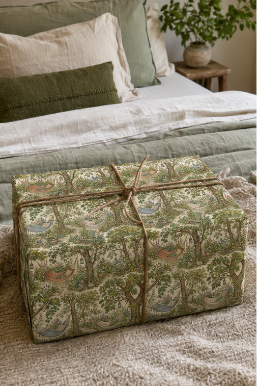 A gift box wrapped in a woodland hammock wrapping paper  with trees, birds, and small houses, tied with a rustic jute twine bow, set on a bed in a cozy bedroom.