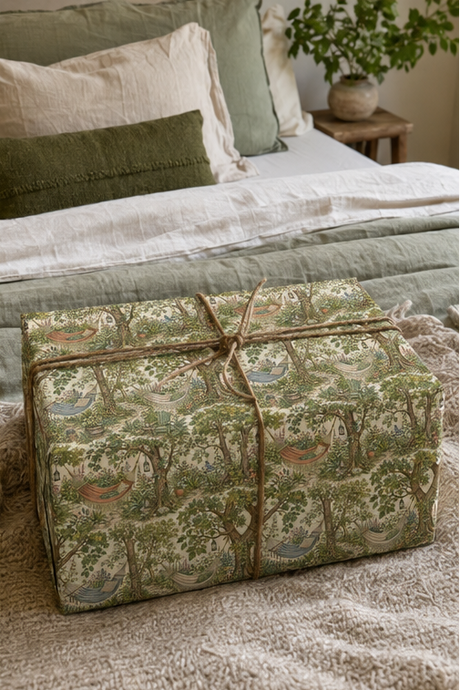 A gift box wrapped in a woodland hammock wrapping paper  with trees, birds, and small houses, tied with a rustic jute twine bow, set on a bed in a cozy bedroom.