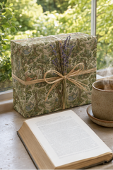 A rectangular gift box wrapped in woodland hammock wrapping paper green and beige botanical-patterned paper with a natural twine bow and dried lavender sprigs, placed on a windowsill beside an open book and a steaming mug with a blurred outdoor garden background.