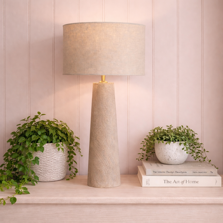 Decorative lamp with a textured base and beige shade on a shelf with books and plants.