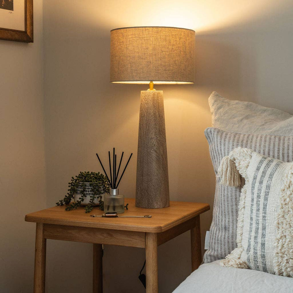 Decorative lamp on a wooden table next to a bed with pillows and a blanket.