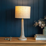 Table lamp with a textured base and beige shade on a wooden surface against a dark blue paneled wall.